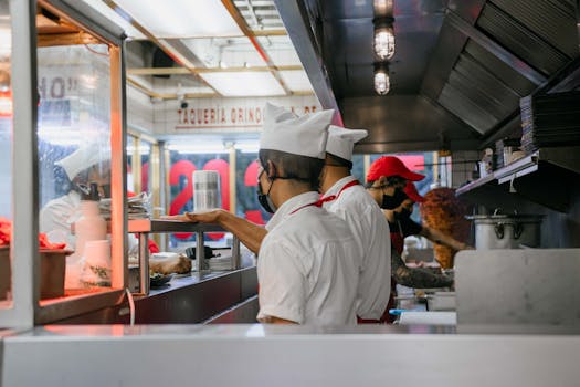 Culinary staff wearing uniforms and masks preparing meals in a bustling restaurant kitchen setting.