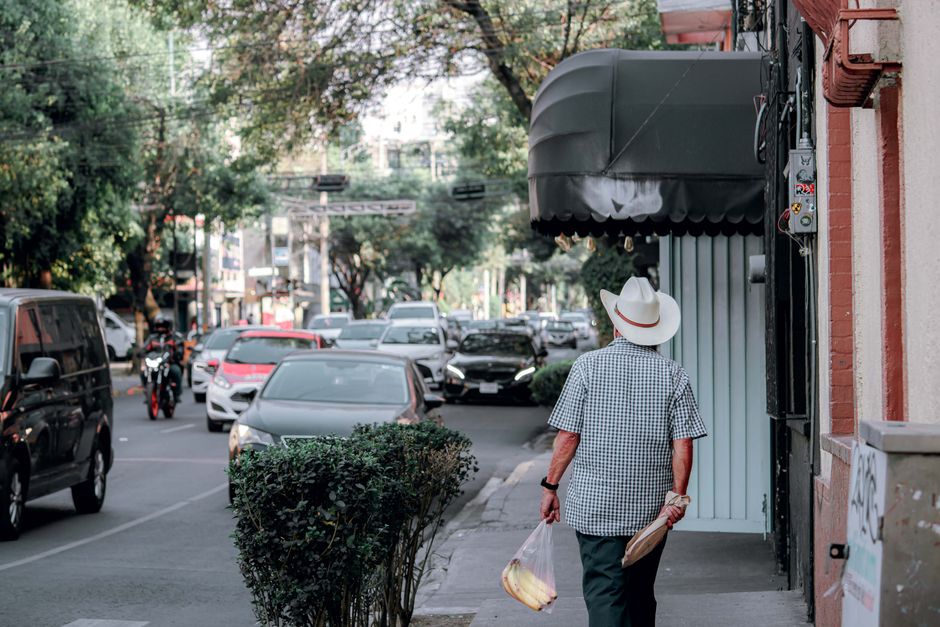 Older man in a fedora walking along a busy city street with groceries in hand.