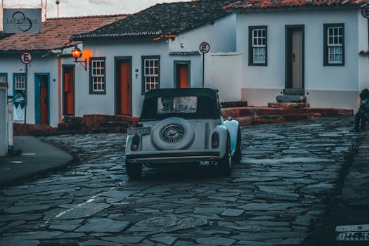 Classic vintage car travels on a cobblestone street lined with historic buildings at dusk.