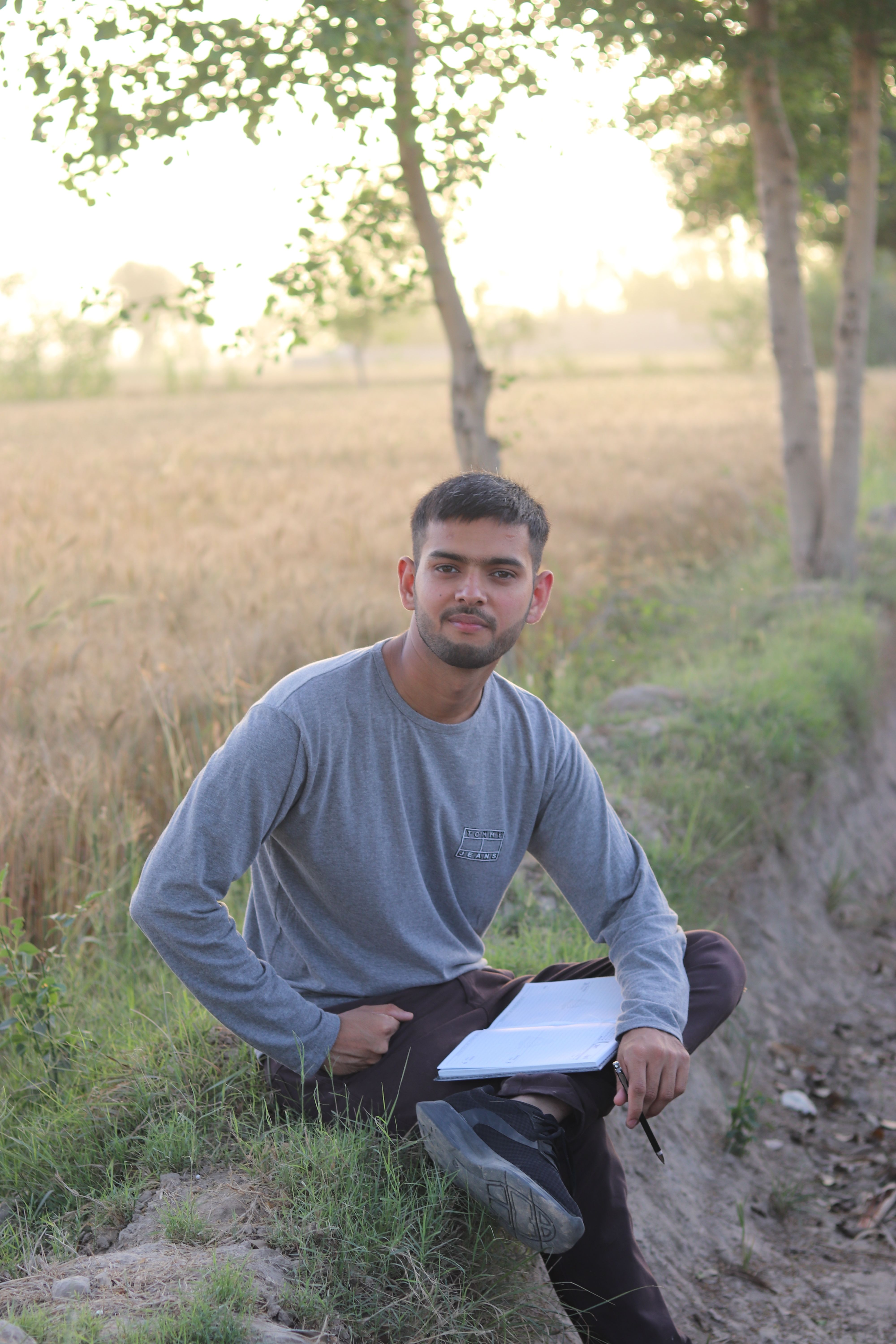 Young man sitting outdoors in a rural setting with trees and fields, enjoying nature.