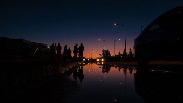Silhouettes of people against a twilight sky with streetlight reflections on water.