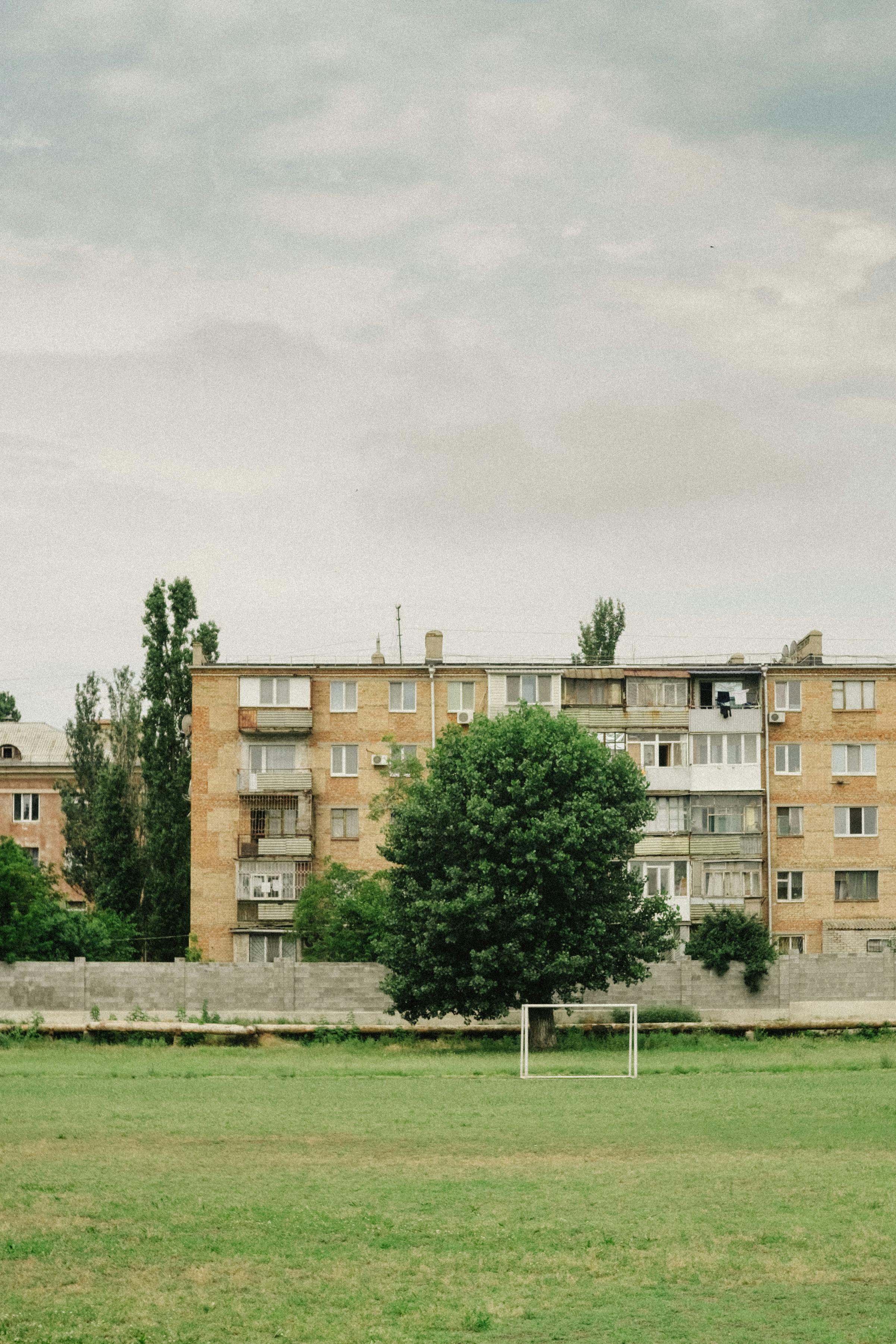 A serene view of an urban soccer field framed by apartment buildings and trees under a cloudy sky.
