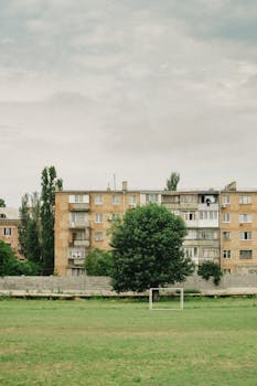 A serene view of an urban soccer field framed by apartment buildings and trees under a cloudy sky.