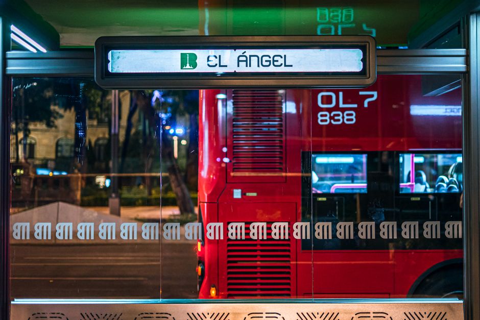 A vibrant night scene of the El Ángel bus stop with a passing red bus in Mexico City.