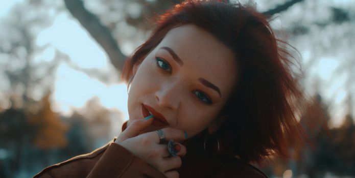 A red-headed woman posing outdoors with red lipstick in a park at sunset.