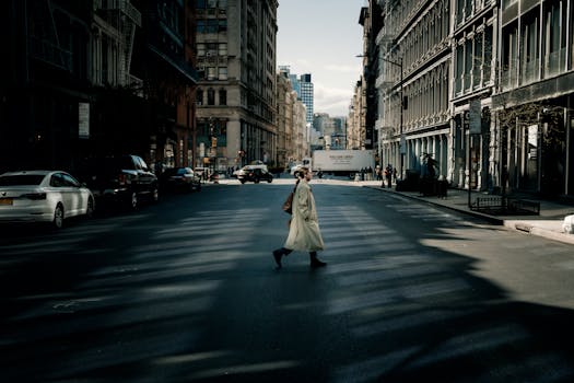 A woman in a trench coat walks across a sunlit street in New York City with tall buildings surrounding her.