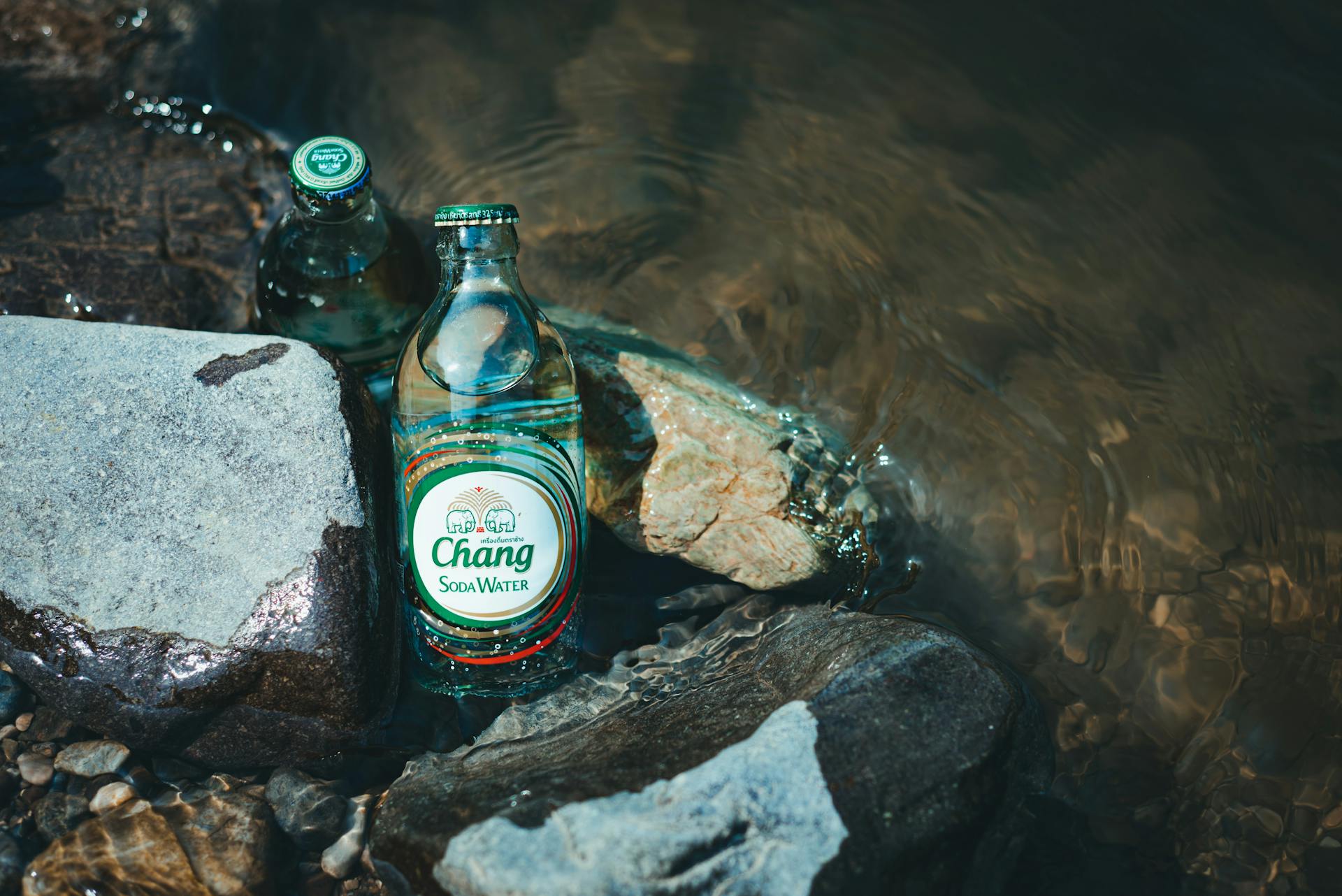 Glass bottles of Chang soda water placed on river rocks with water flowing, creating a refreshing scene.