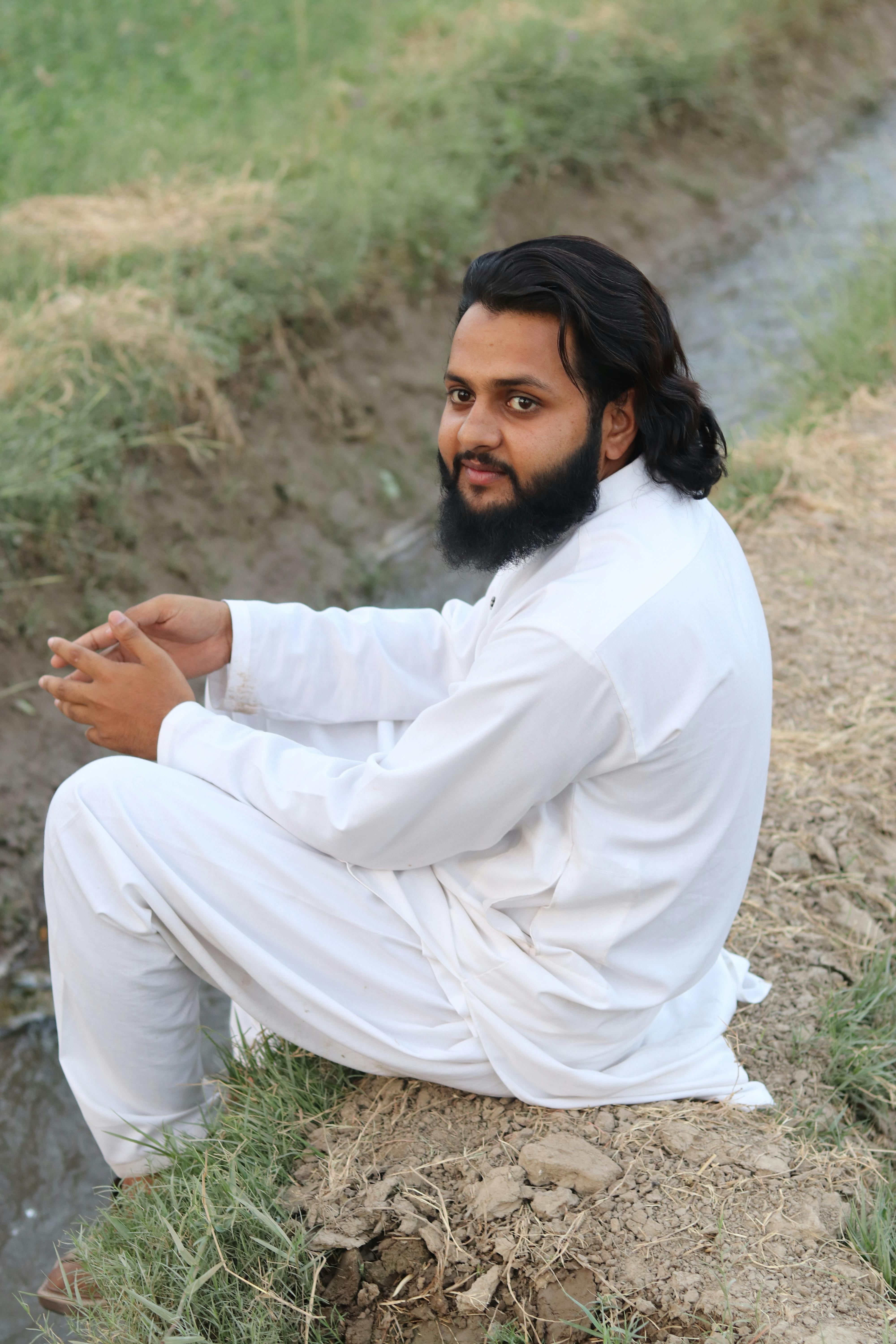 Bearded man in white traditional attire sitting by a field, showcasing cultural style.