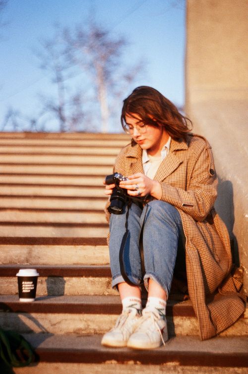 Free A young woman sits on urban stairs holding a camera, enjoying a coffee outdoors. Stock Photo