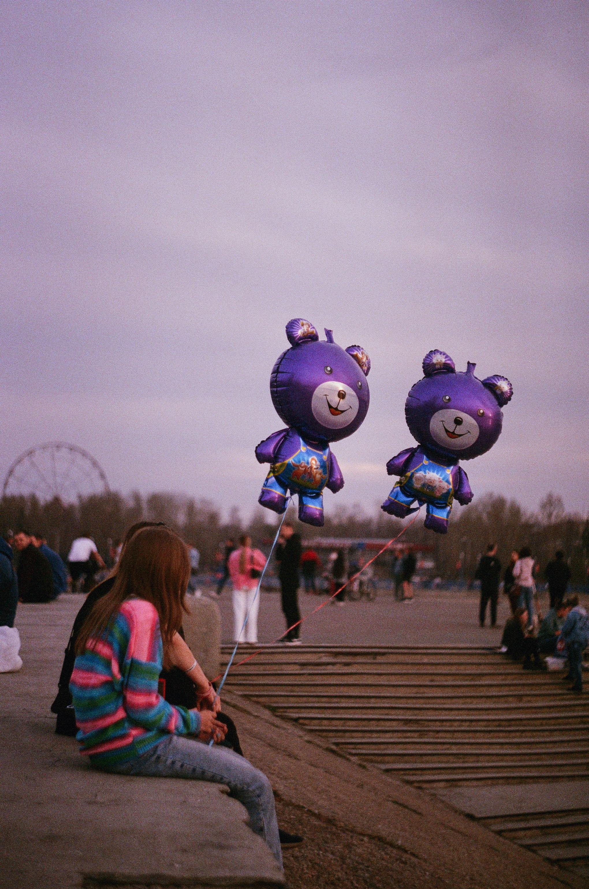 A serene outdoor scene featuring colorful bear balloons and people relaxing in a park at dusk.