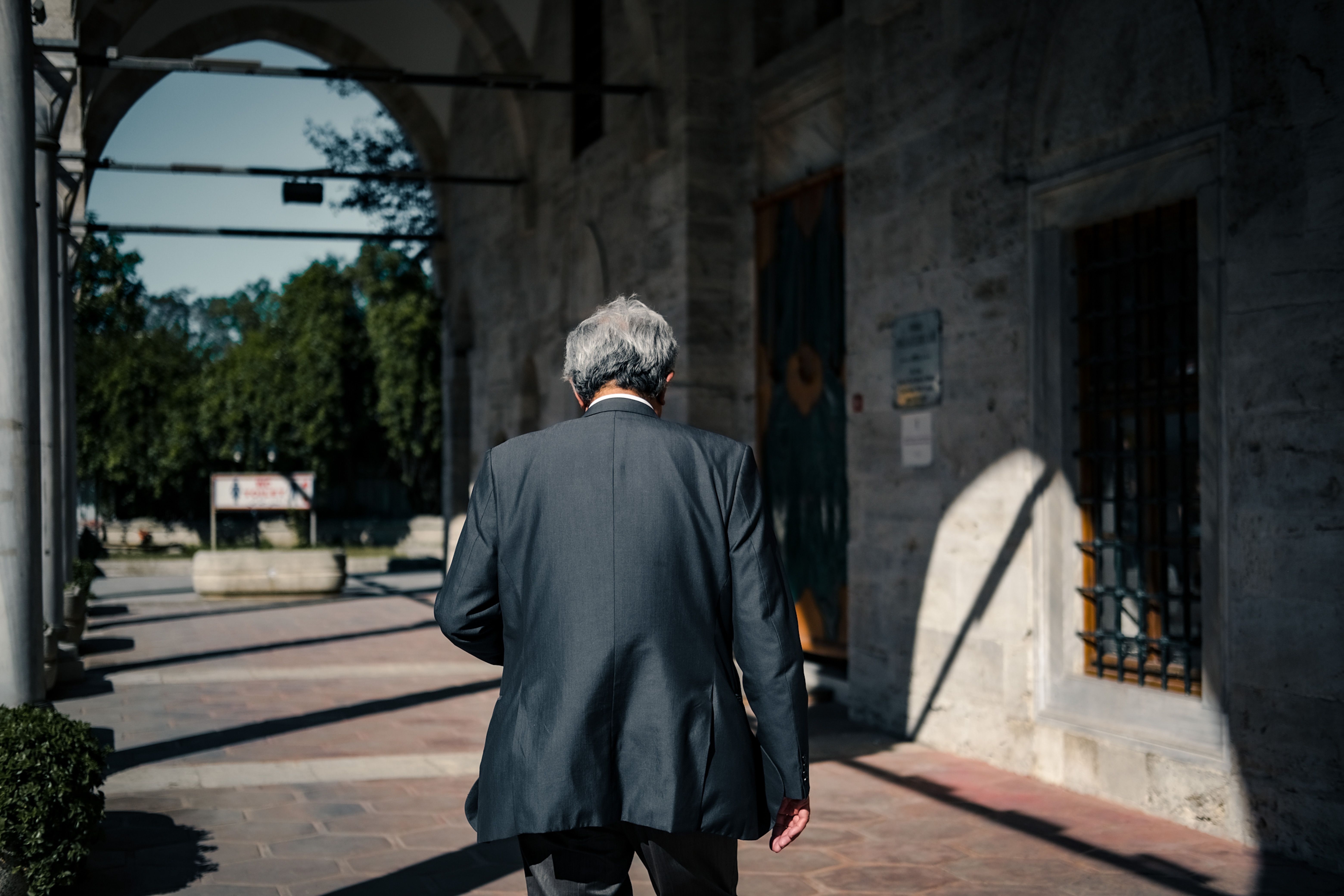 Back view of an elderly man in a suit walking through a sunlit urban corridor, creating dramatic shadows.