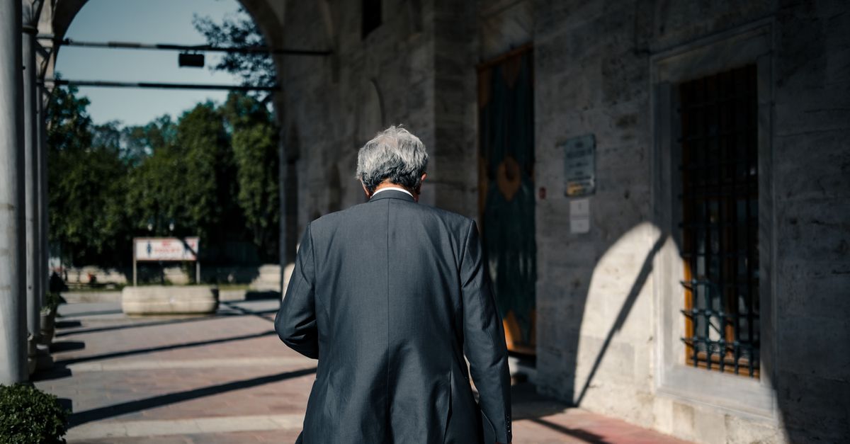 Back view of an elderly man in a suit walking through a sunlit urban corridor, creating dramatic shadows.