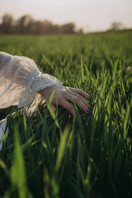 A delicate hand with silver rings gently touches the green grass under warm sunlight, conveying serenity.