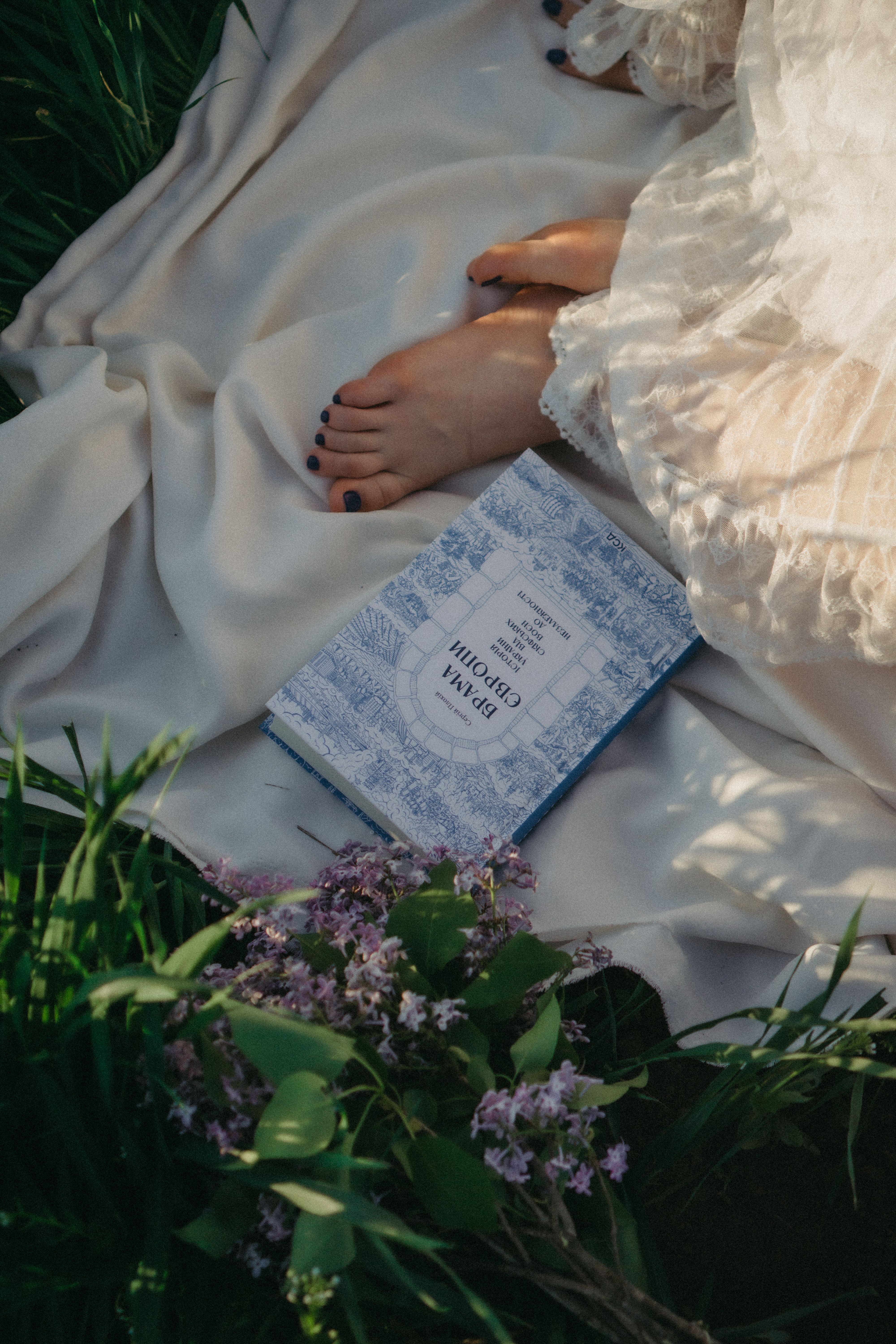 A woman relaxes barefoot on a blanket outdoors, reading a book amidst flowers in the grass.