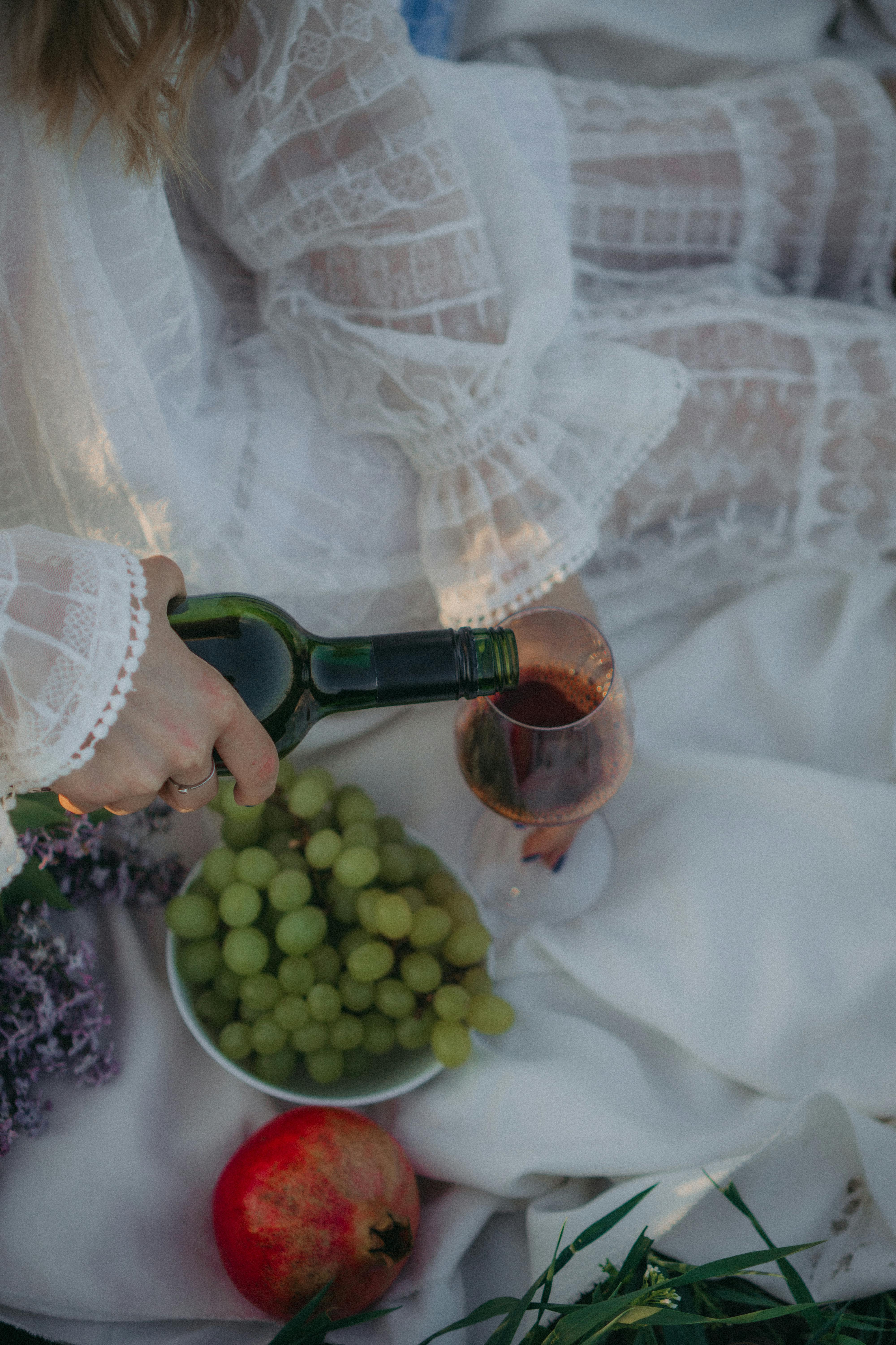 A serene picnic setup featuring wine pouring, grapes, and a pomegranate on white fabric.