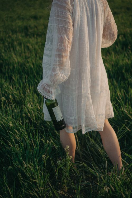 Free A woman in a white dress holds a bottle while standing in a grassy field during the day. Stock Photo