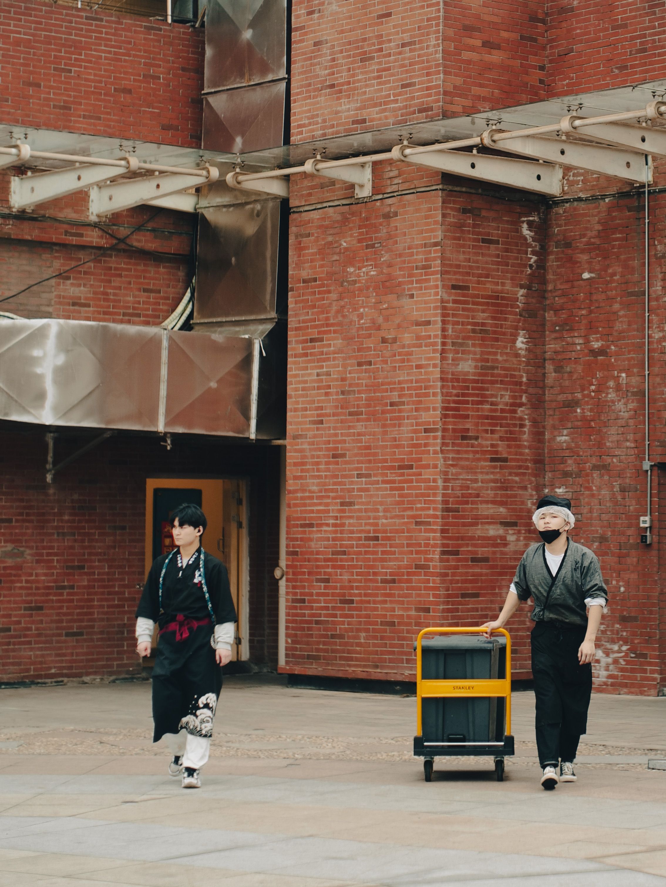 Two men in traditional attire walk past an urban brick building with luggage, showcasing cultural diversity.