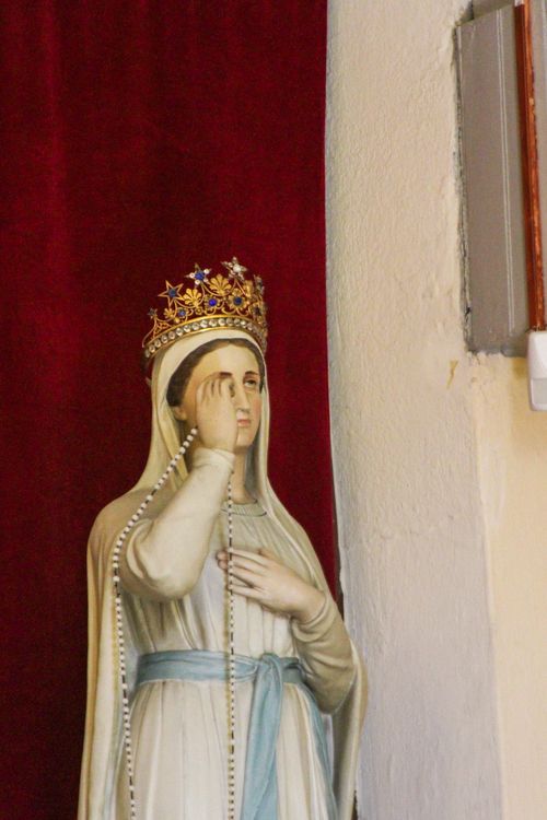 Free Statue of Virgin Mary with crown and rosary against a red curtain backdrop. Stock Photo