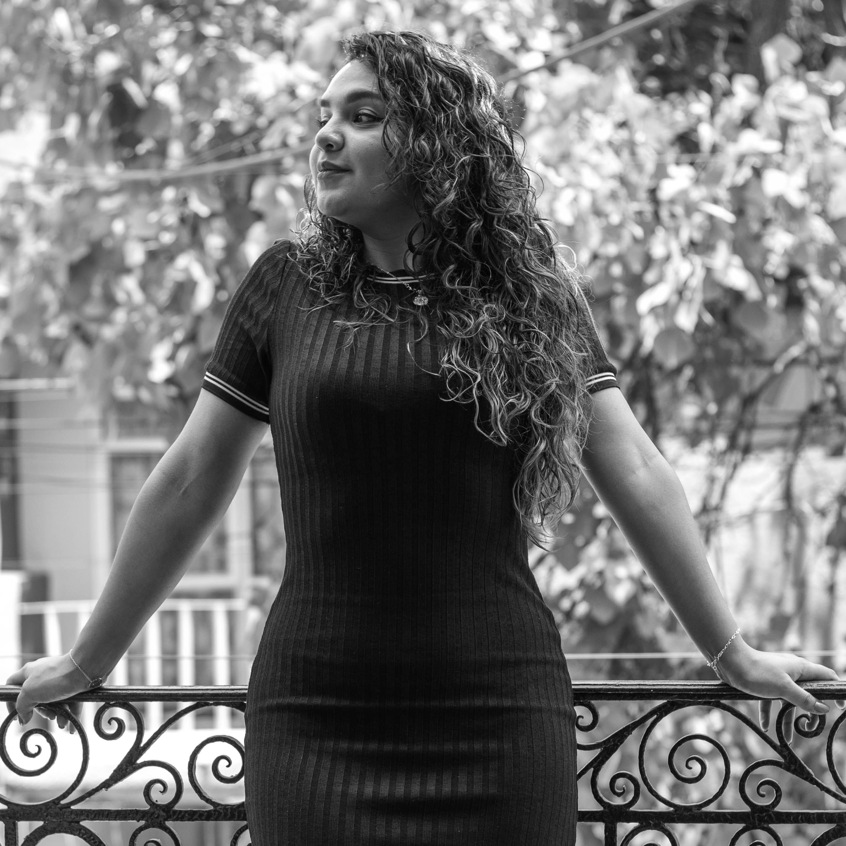 Confident woman in a black dress poses gracefully on a balcony, embracing city vibes.