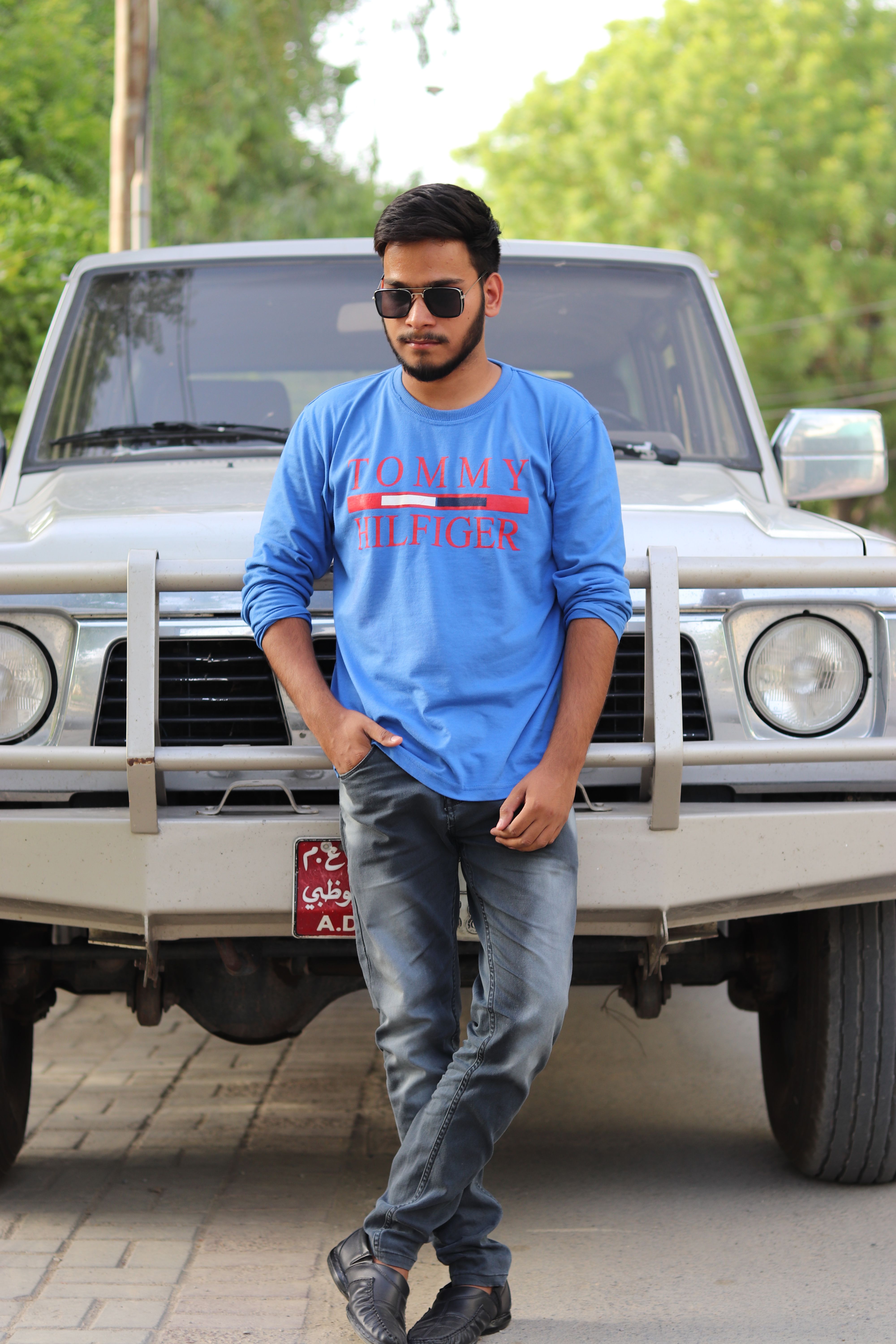 Man in blue long sleeves with sunglasses leaning on a white SUV outdoors.