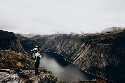 Free Person standing on a cliff edge overlooking a dramatic fjord landscape in Norway under overcast skies. Stock Photo