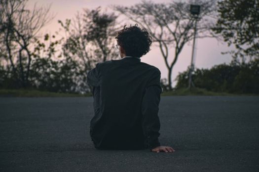 A man with curly hair sits on a road in solitude at dusk, offering a contemplative scene.