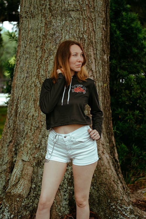 A Woman in Black Sweater and Denim Shorts Standing Beside Brown Tree