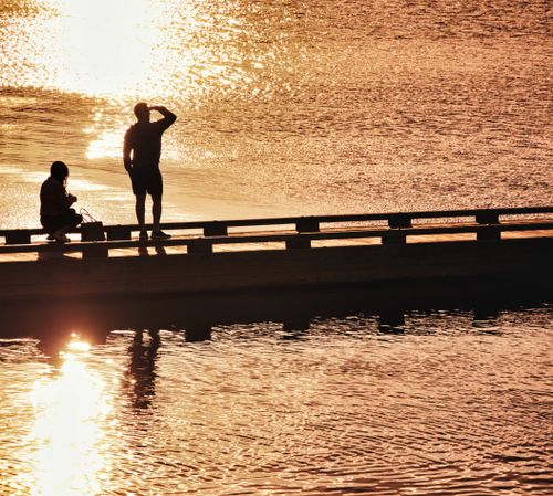 Free A couple silhouetted against a golden sunset, standing on a pier overlooking shimmering water. Stock Photo