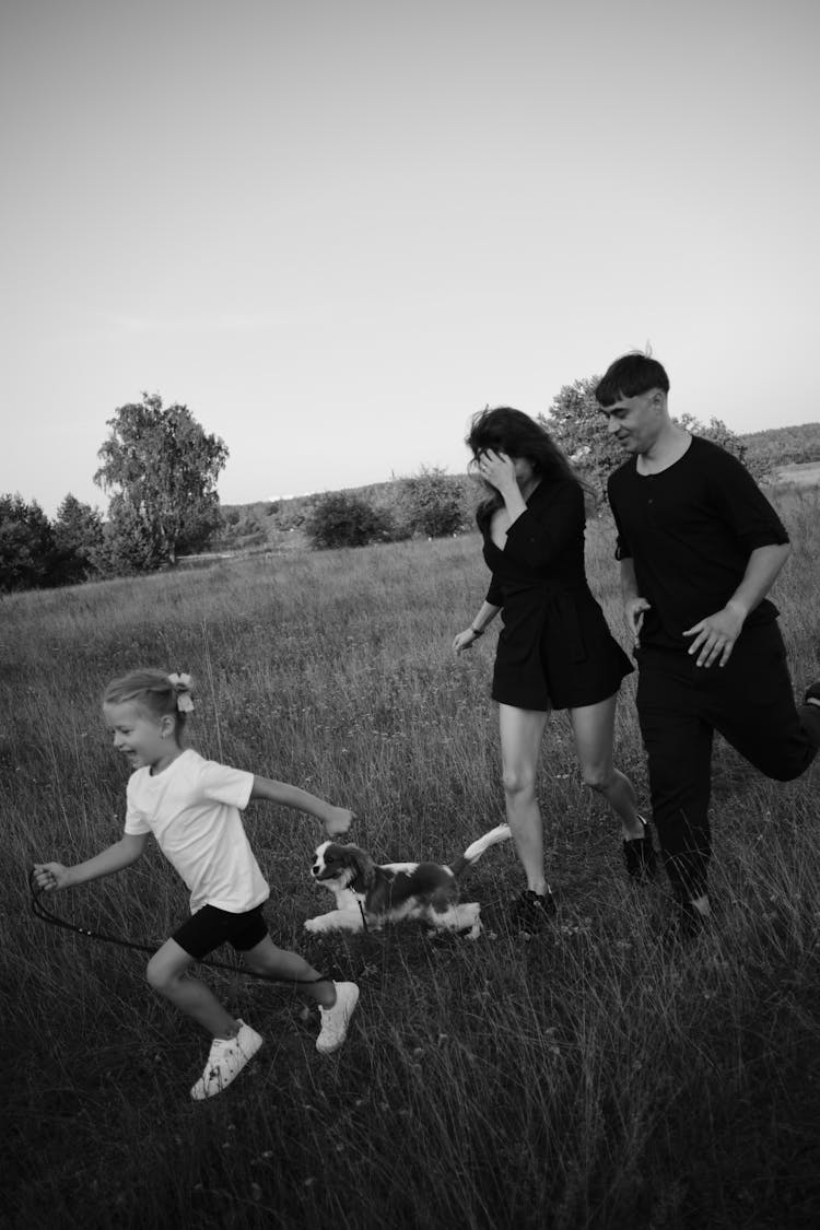 Little Girl With Parents And Dog Running On A Field 