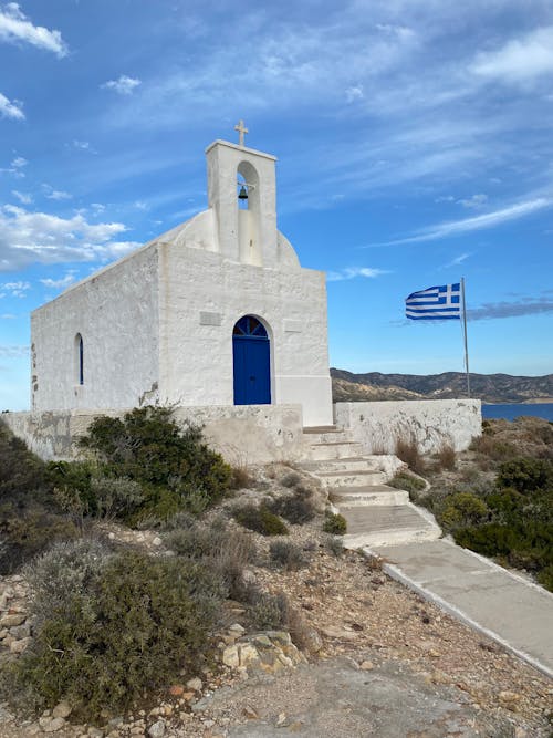 Free White Concrete Chapel with Blue Door Near Flag of Greece Stock Photo