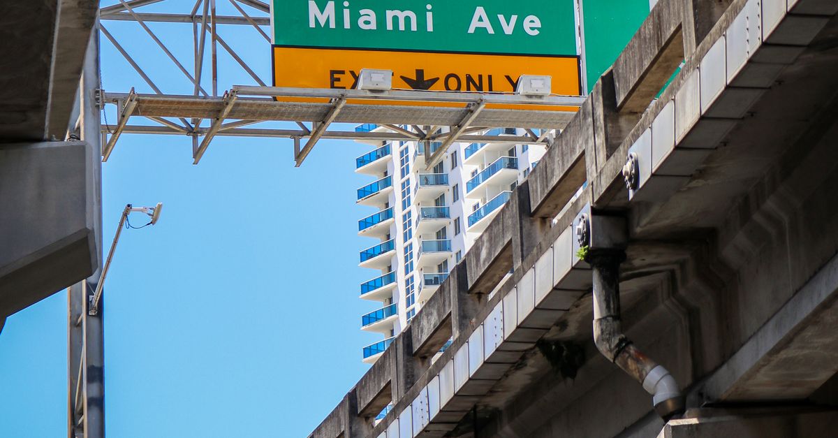 Elevated road sign for Downtown Miami Ave with skyscrapers in background, blue sky above.