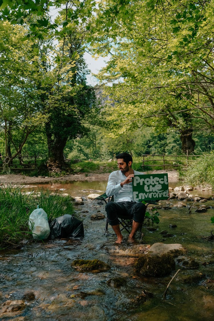 Bearded Man In White Dress Shirt Sitting On Chair In The River Holding A Sign