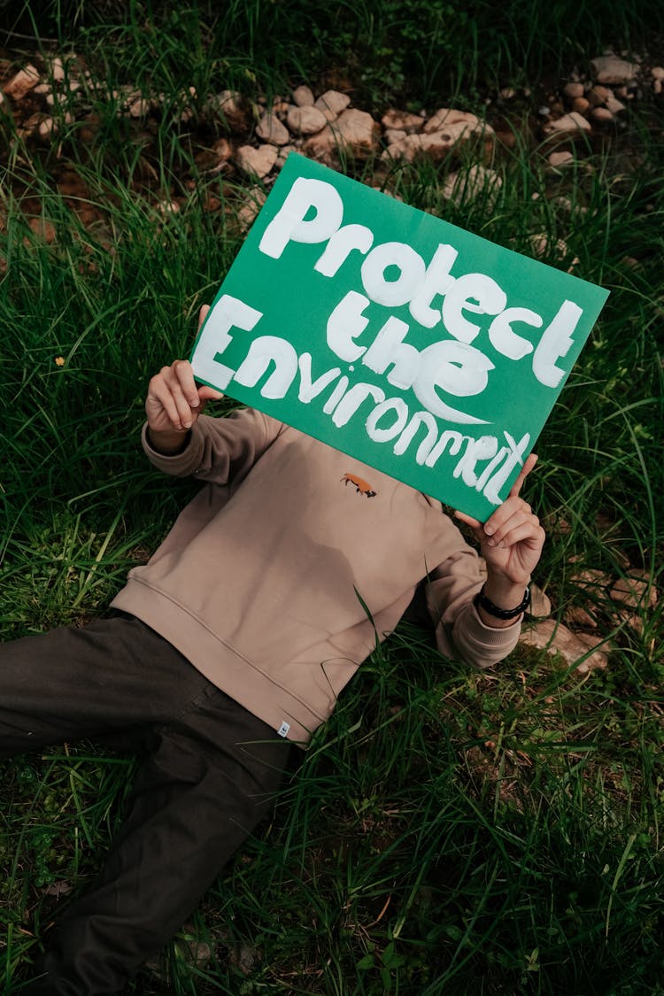 A Person Lying On Green Grass Holding A Protect The Environment Placard
