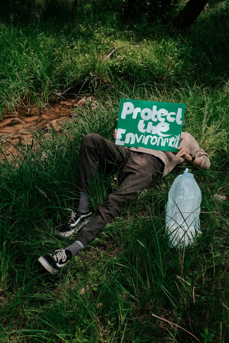 A Person Lying On Green Grass Holding A Protect The Environment Placard
