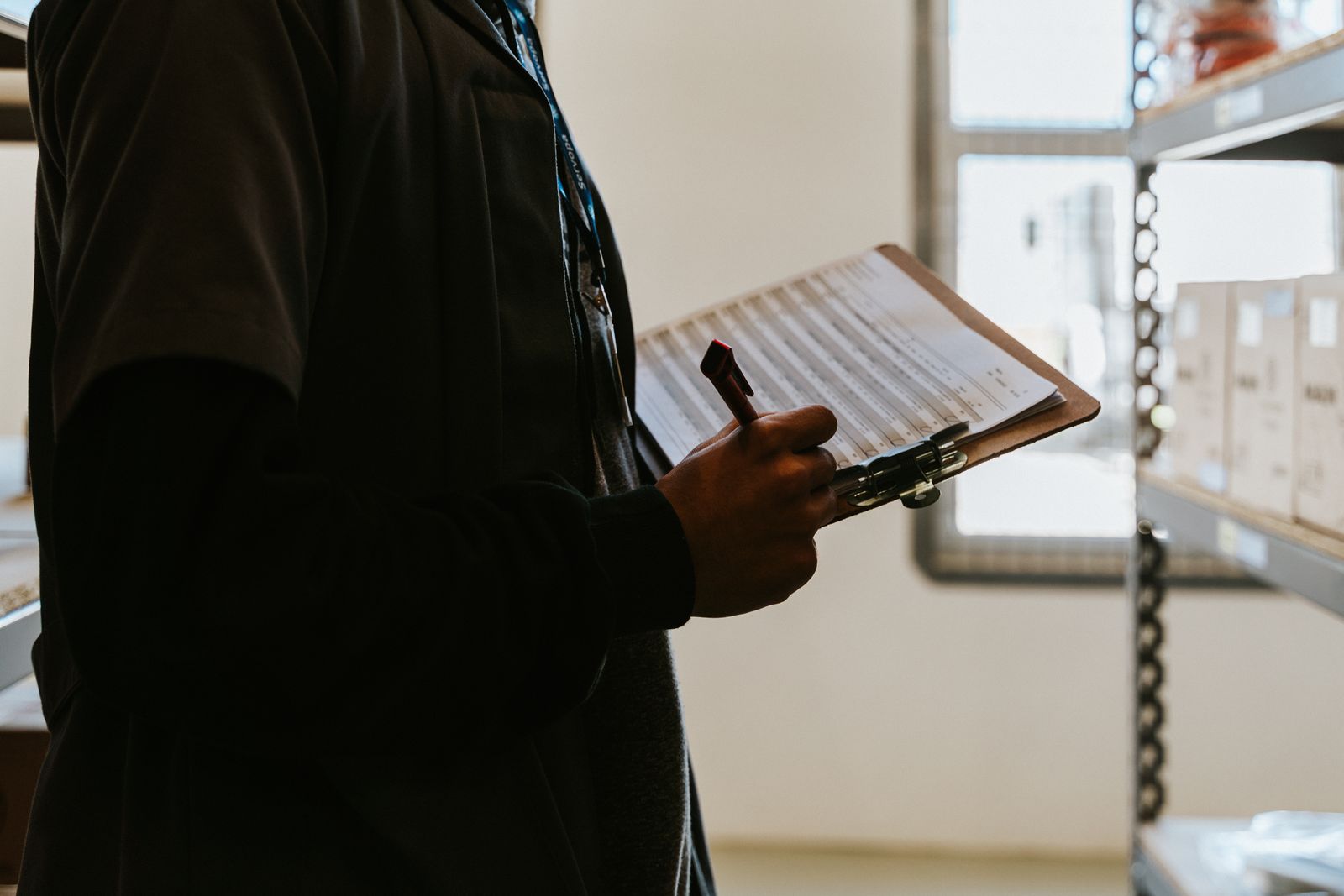 Warehouse staff member checking stock against a printed checklist on a clipboard beside storage shelves