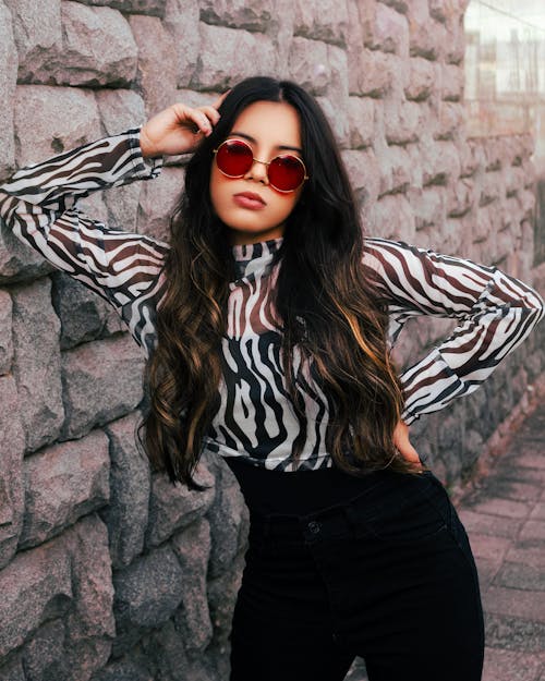 A Woman in Printed Long Sleeves Wearing Sunglasses while Posing Near the Stone Wall