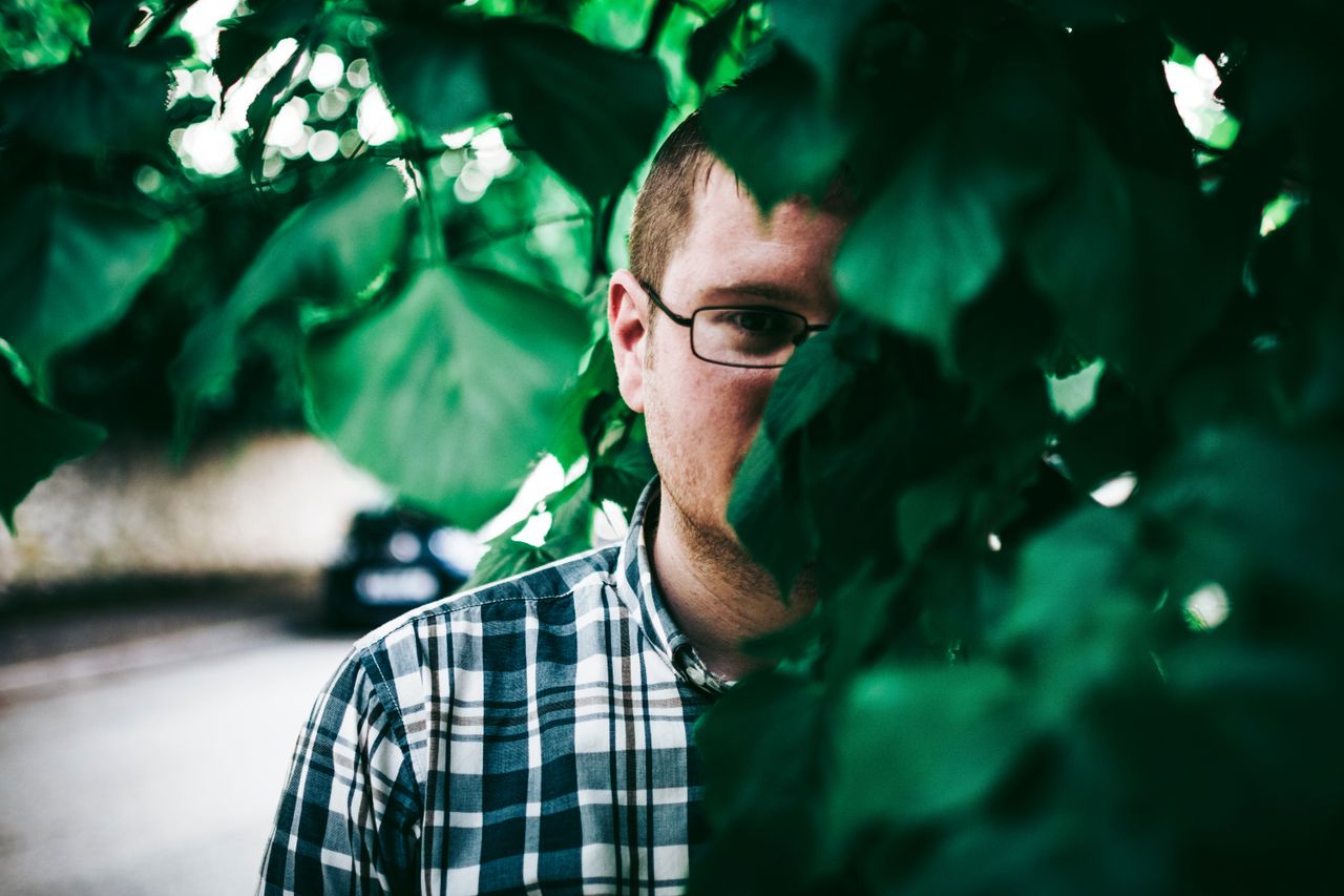 a man wearing glasses partially obscured by greenery outdoors creating