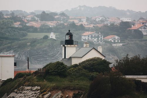 Free Charming coastal lighthouse surrounded by lush hills and red-roofed homes in a misty seaside environment. Stock Photo