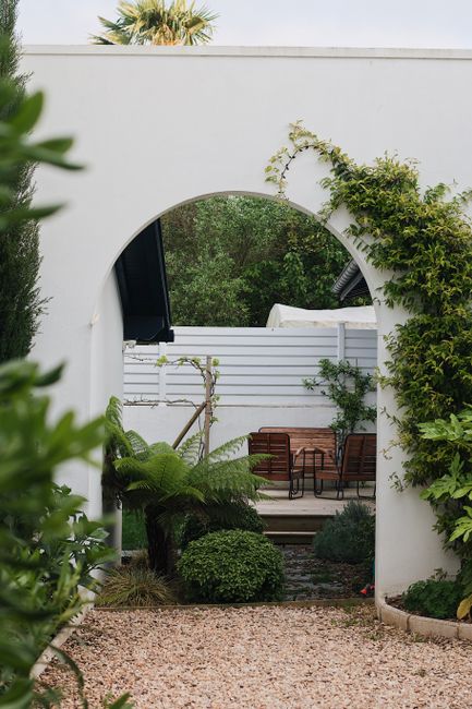 Photo by Maël BALLAND on Pexels A serene garden view through a modern arch surrounded by lush greenery and patio chairs.