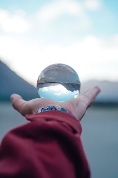 A crystal ball in a person's hand reflects a calm landscape with mountains and sky.