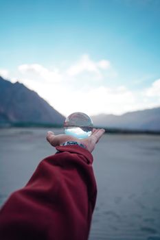 A hand holding a glass ball reflecting mountains with a blurred natural landscape background.
