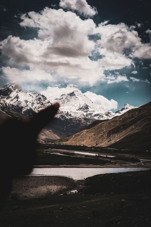Free Scenic view of snow-capped mountains and river under dramatic sky, featuring a pointing silhouette in the foreground. Stock Photo