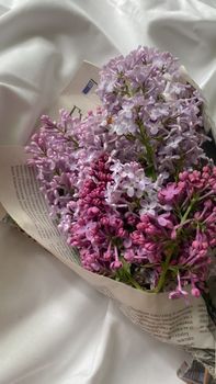 Capture of a delicate lilac bouquet wrapped in newspaper on a soft fabric background.