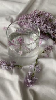 Serene arrangement of lilac flowers delicately placed in a clear glass of water on white fabric.