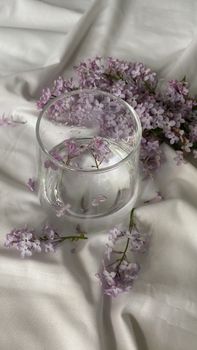 A serene still life of lilac blossoms arranged around a clear glass on white fabric, captured in natural light.