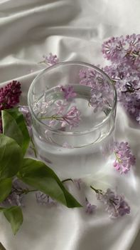 A glass of water beautifully adorned with delicate lilac flowers on a silky white background.
