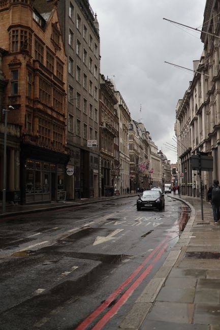 A rainy city street with historic buildings and a moving car under a cloudy sky.