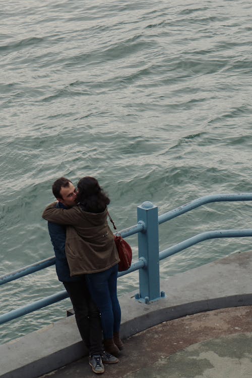 Woman in Green Jacket Hugging Man in Blue Jacket Near Body of Water