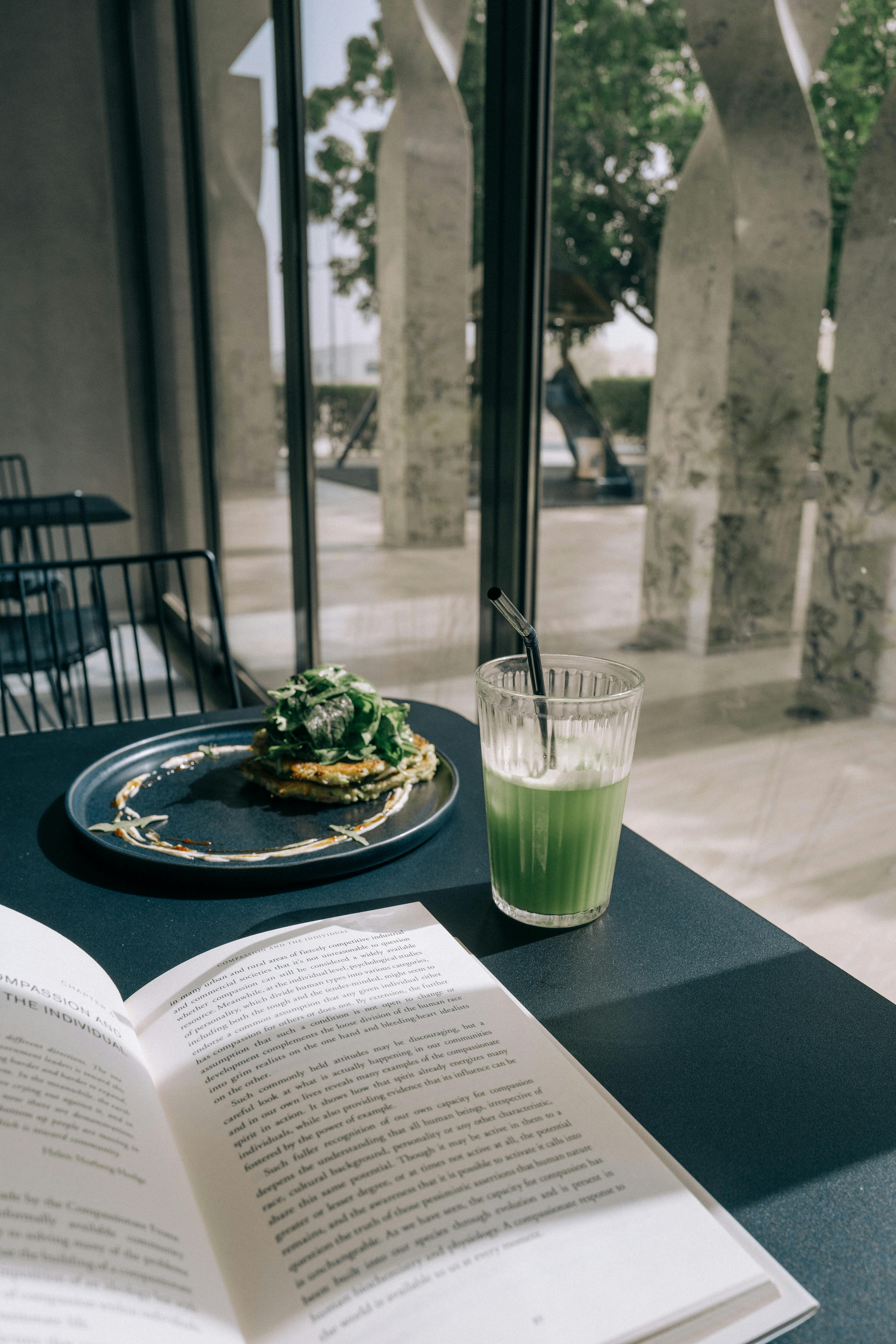 Open book next to a green smoothie and plate of food on table by a sunny window.