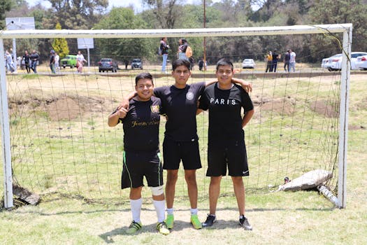 Three young men wearing sportswear stand smiling at a soccer goal on a sunny day.